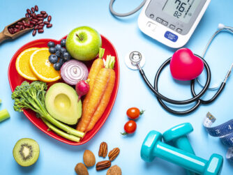 Healthy lifestyle concepts: red heart shape plate with fresh organic fruits and vegetables shot on blue background. A digital blood pressure monitor, doctor stethoscope, dumbbells and tape measure are beside the plate This type of foods are rich in antioxidants and flavonoids that prevents heart diseases, lower cholesterol and help to keep a well balanced diet.