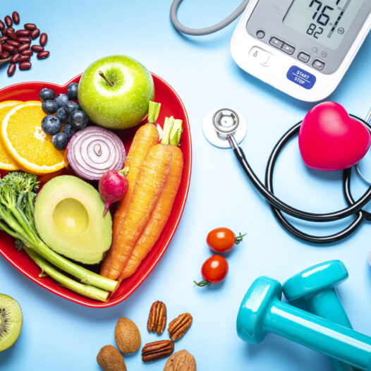 Healthy lifestyle concepts: red heart shape plate with fresh organic fruits and vegetables shot on blue background. A digital blood pressure monitor, doctor stethoscope, dumbbells and tape measure are beside the plate This type of foods are rich in antioxidants and flavonoids that prevents heart diseases, lower cholesterol and help to keep a well balanced diet.