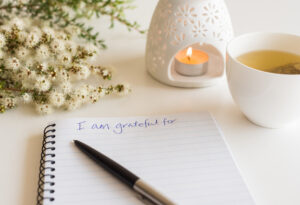 Close up of handwritten text "I am grateful for..." in foreground with notebook, pen, cup of tea, flowers and oil burner in soft focus