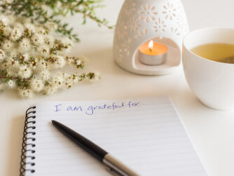 Close up of handwritten text "I am grateful for..." in foreground with notebook, pen, cup of tea, flowers and oil burner in soft focus