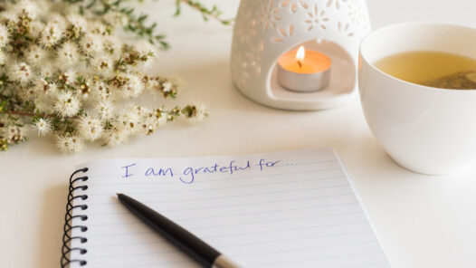 Close up of handwritten text "I am grateful for..." in foreground with notebook, pen, cup of tea, flowers and oil burner in soft focus