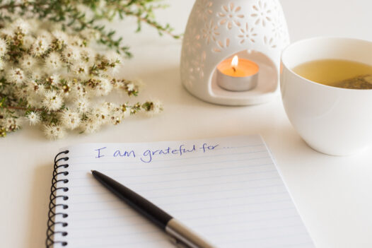 Close up of handwritten text "I am grateful for..." in foreground with notebook, pen, cup of tea, flowers and oil burner in soft focus