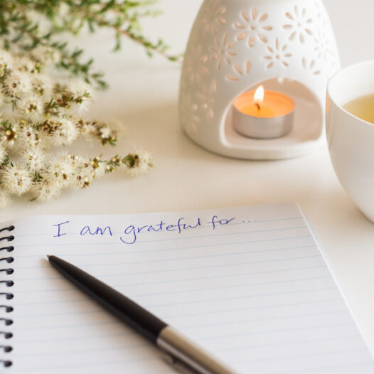 Close up of handwritten text "I am grateful for..." in foreground with notebook, pen, cup of tea, flowers and oil burner in soft focus