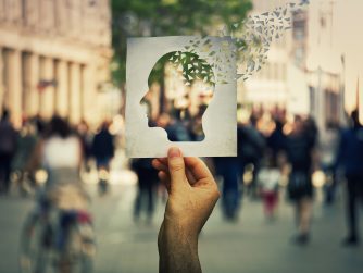 Hand holding a paper sheet with human head icon broken into pieces over a crowded street background. Concept of memory loss and dementia disease. Alzheimer's losing brain and memory function.