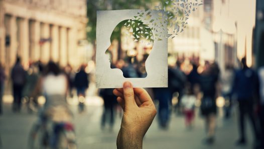 Hand holding a paper sheet with human head icon broken into pieces over a crowded street background. Concept of memory loss and dementia disease. Alzheimer's losing brain and memory function.