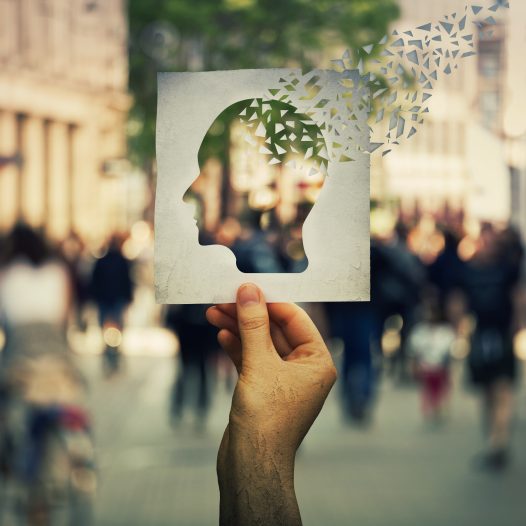 Hand holding a paper sheet with human head icon broken into pieces over a crowded street background. Concept of memory loss and dementia disease. Alzheimer's losing brain and memory function.