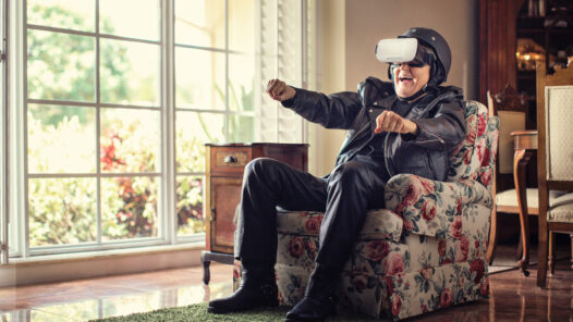 Old man sitting in an arm chair wearing a leather jacket, motorcycle helmet and VR headset.