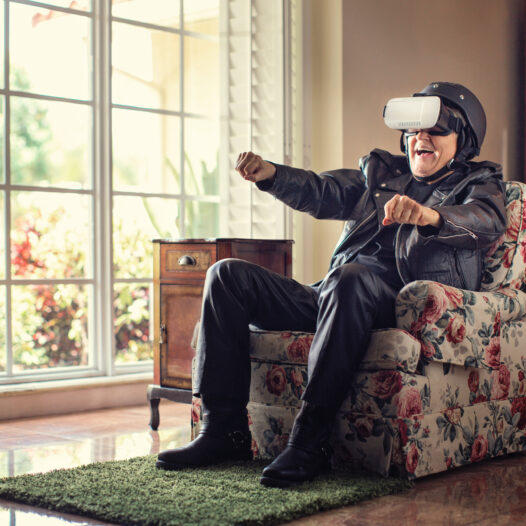 Old man sitting in an arm chair wearing a leather jacket, motorcycle helmet and VR headset.