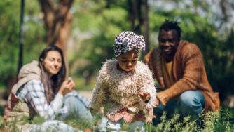 Inter-racial family at a park. Mother and father watching toddler pick flowers.