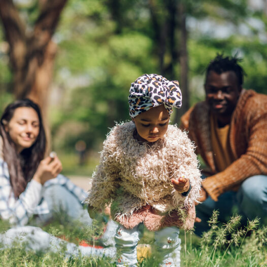 Inter-racial family at a park. Mother and father watching toddler pick flowers.